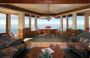 A cozy room with a wooden ceiling, large windows offering a view of the ocean, a round table with chairs, and two sofas with patterned cushions.
