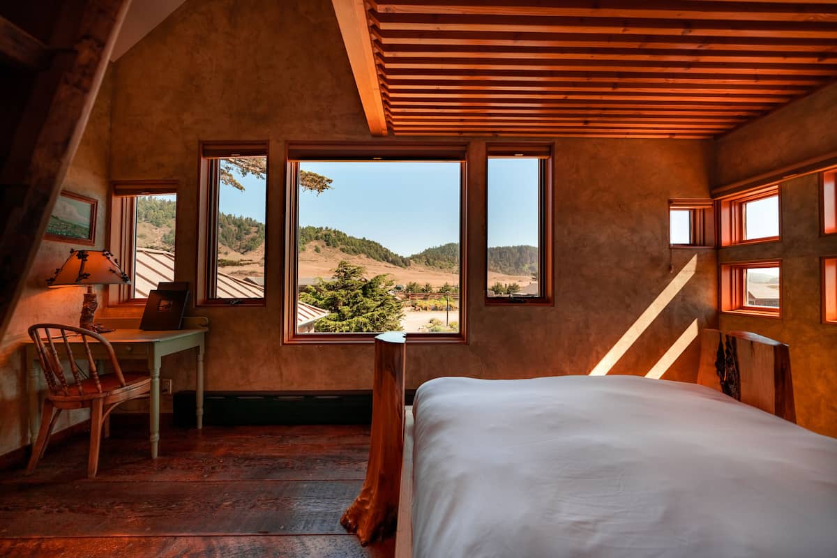Un dormitorio con cama, escritorio y silla, amplias ventanas con vistas a un paisaje montañoso.
