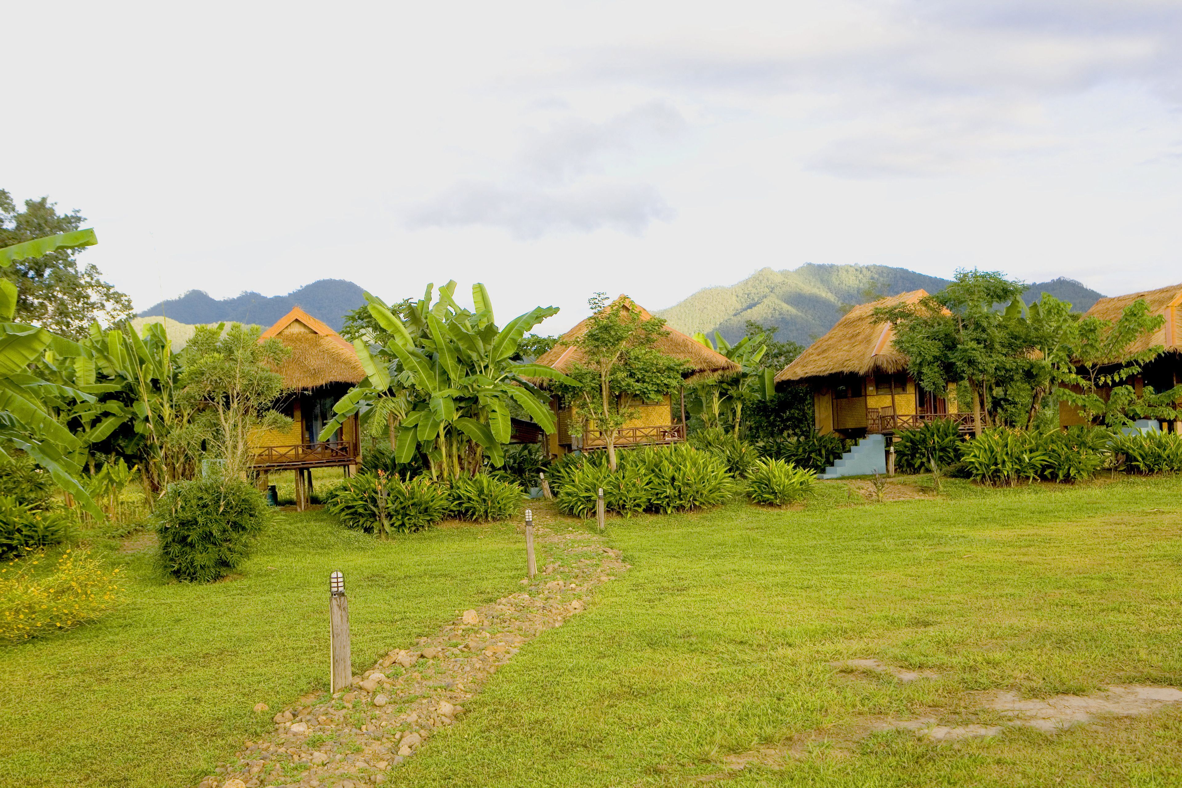 A row of thatched-roof huts with a grassy path leading to them, surrounded by lush greenery and mountains in the background.