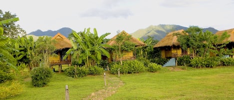 A row of thatched-roof huts with a grassy path leading to them, surrounded by lush greenery and mountains in the background.