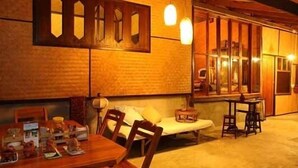 A cozy room with a wooden dining table, chairs, and a sofa. There are two windows with curtains, and a small table with a lamp.