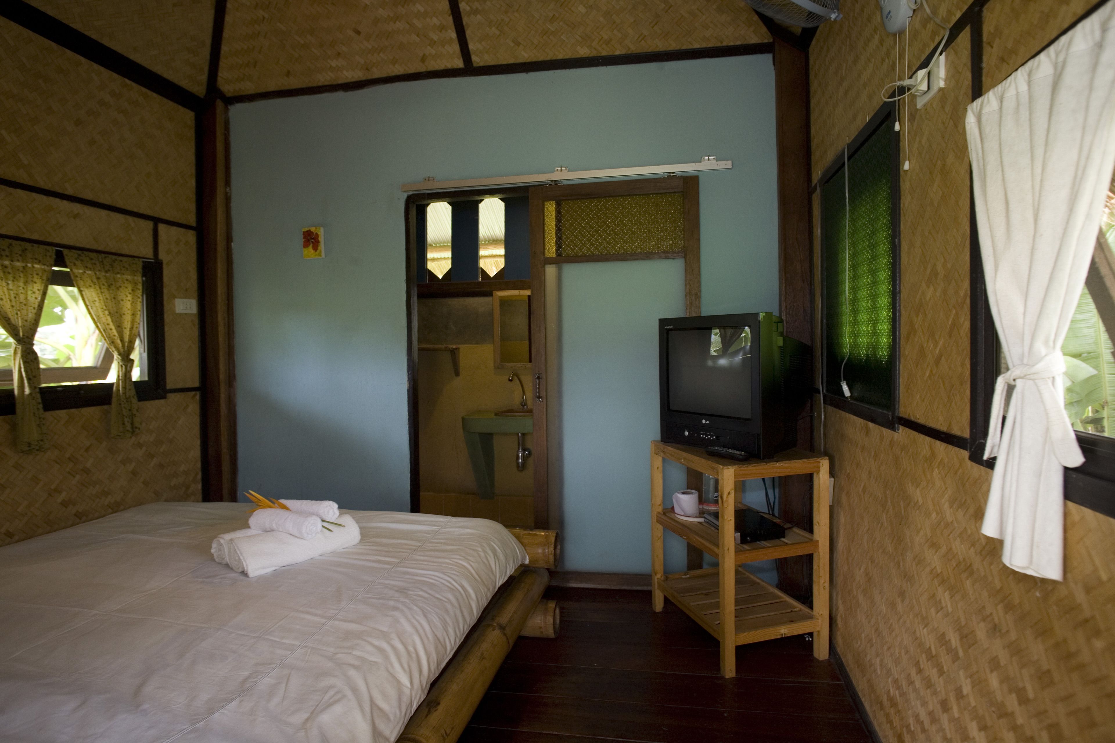 A room with a bed, a television on a wooden stand, a bathroom visible through a doorway, and a window with curtains.