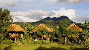 A row of thatched-roof huts with a lush garden in front, set against a backdrop of a mountain and partly cloudy sky.