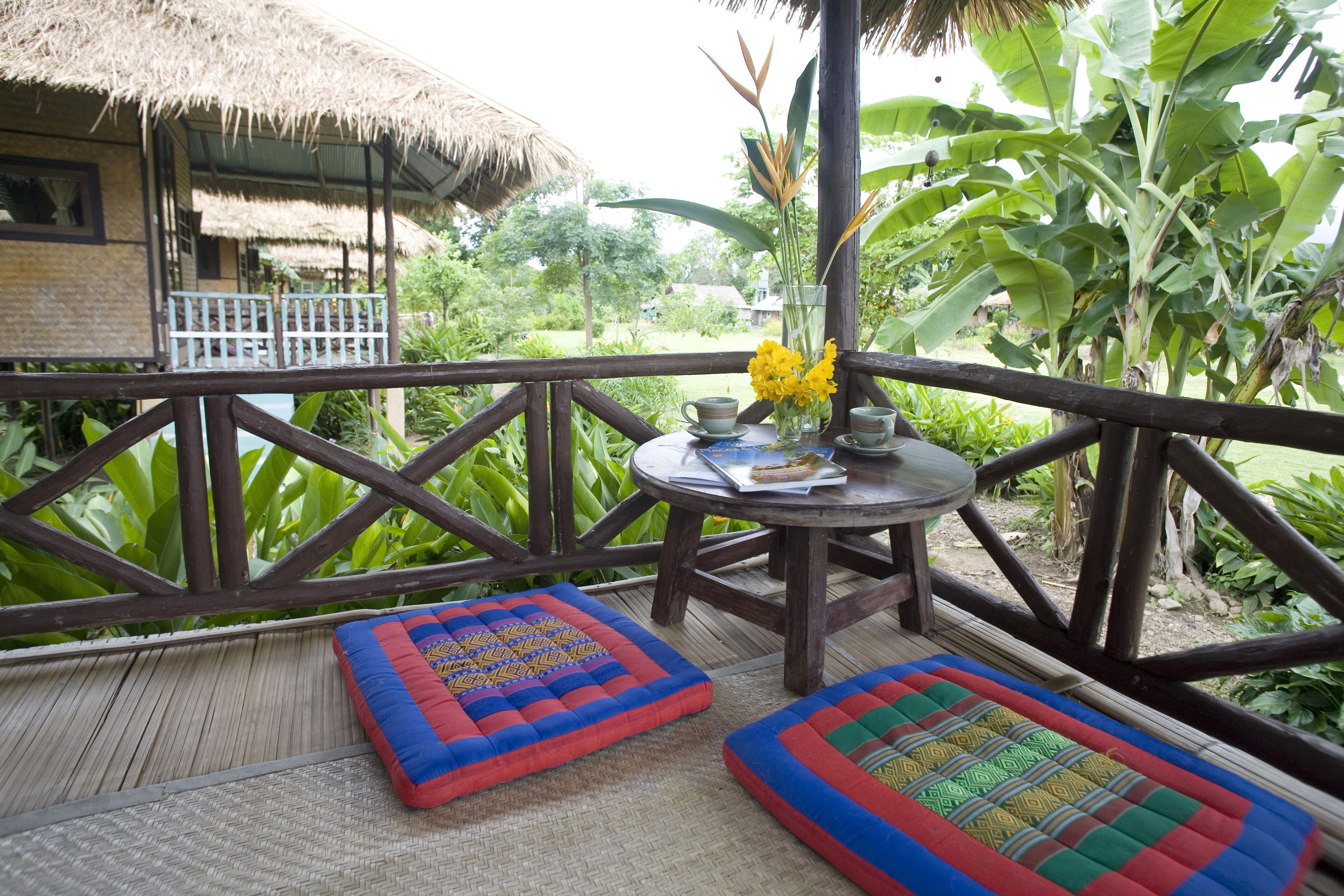 A wooden deck with a thatched roof structure, a round table with a vase of yellow flowers, and two colorful cushions.
