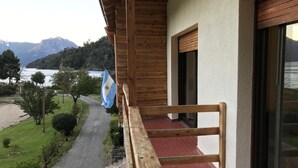 A wooden balcony with a flag, a pathway leading to a beach, and a view of a lake and mountains. - Alun Nehuen (San Carlos de Bariloche)