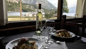 A dining table with plates of food, wine glasses, and a bottle of wine. A window reveals a mountainous landscape and a body of water. - Alun Nehuen (San Carlos de Bariloche)