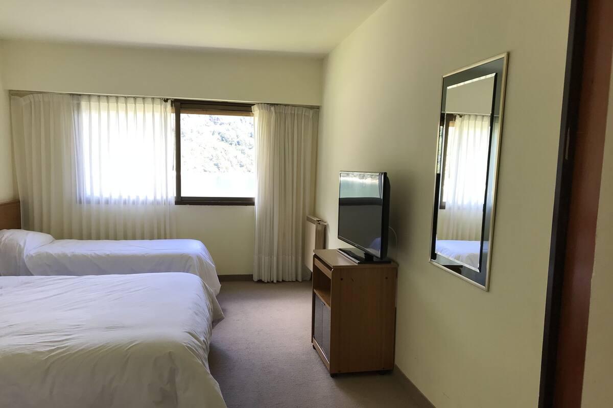 Une chambre d’hôtel avec deux lits, un téléviseur posé sur un meuble en bois et une fenêtre avec des rideaux.