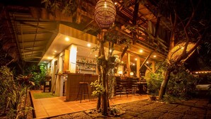 Zona de bar a l'exterior amb sostre de fusta rústica, envoltada de vegetació tropical exuberància i un camí empedrat.