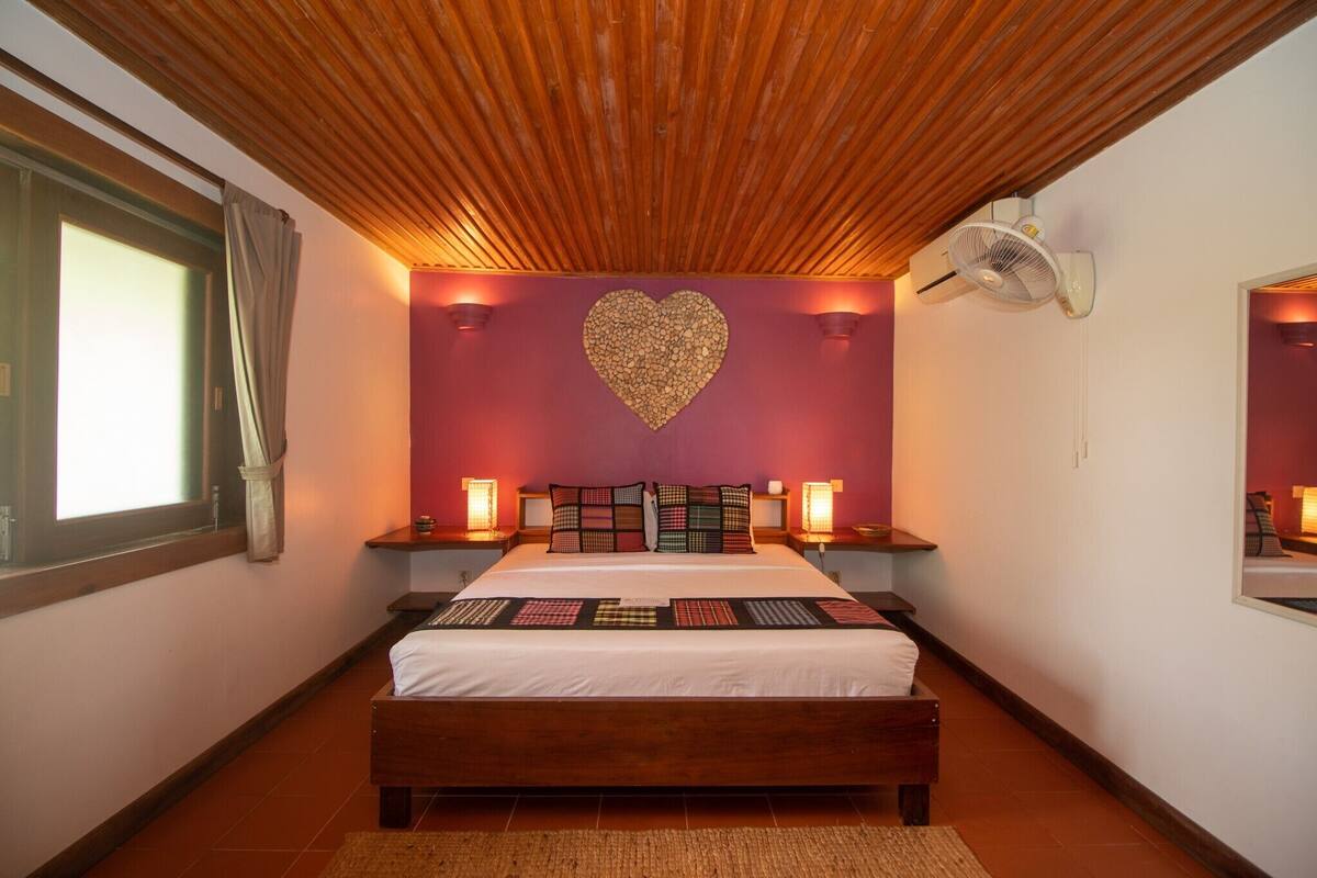 A bedroom with a wooden ceiling, a heart-shaped wall decoration, a bed with patterned bedding, bedside lamps, and a wall-mounted fan.