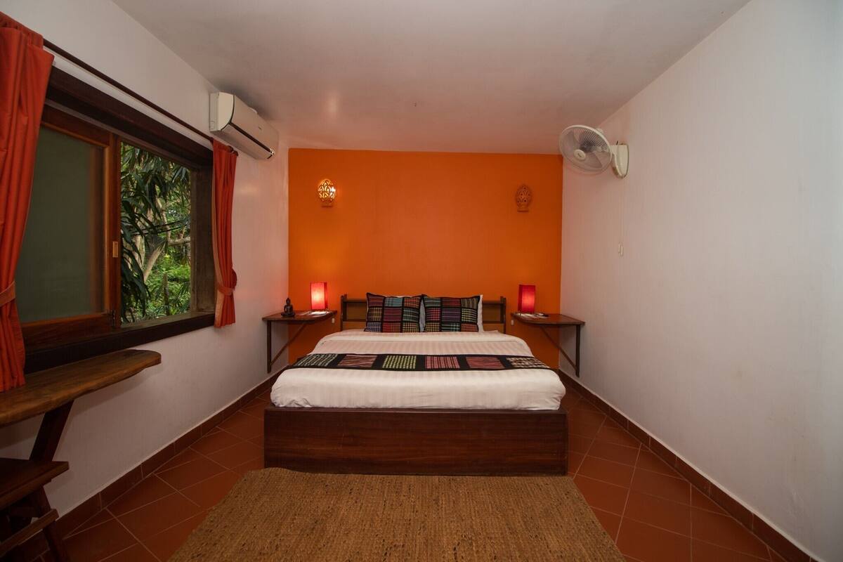 A bedroom with a bed, two bedside tables, a nightstand, a window with curtains, an air conditioner, a wall-mounted fan, and a view of greenery outside.