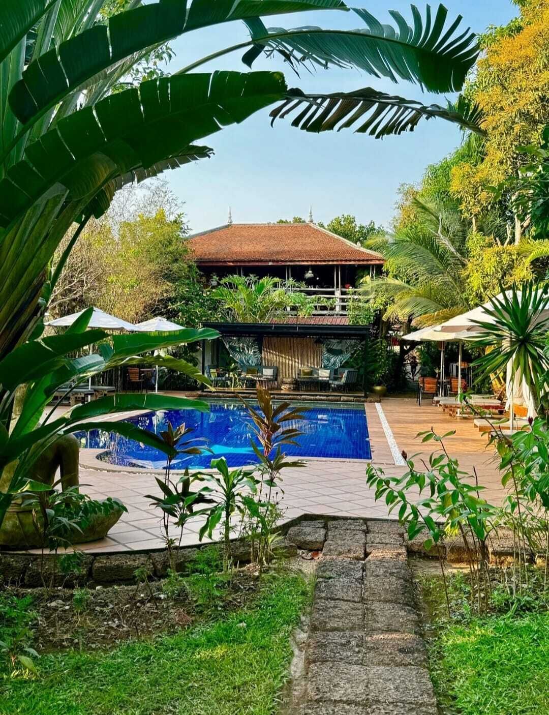 Área de piscina com vista clara para um edifício com telhado em azulejos, rodeada por vegetação exuberante e mobiliário de exterior.