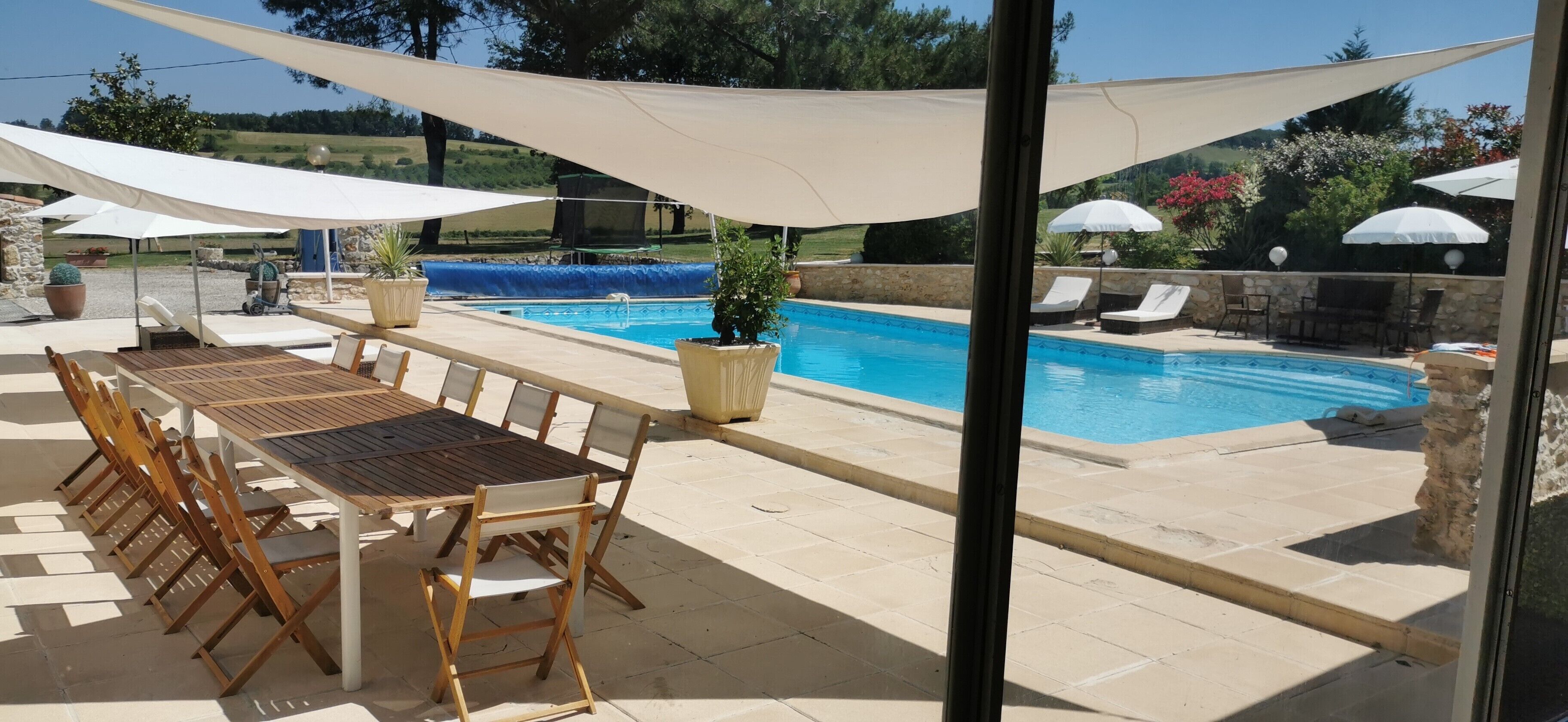 Zona junto a la piscina con mesa y sillas de madera, sombrillas y plantas en macetas.