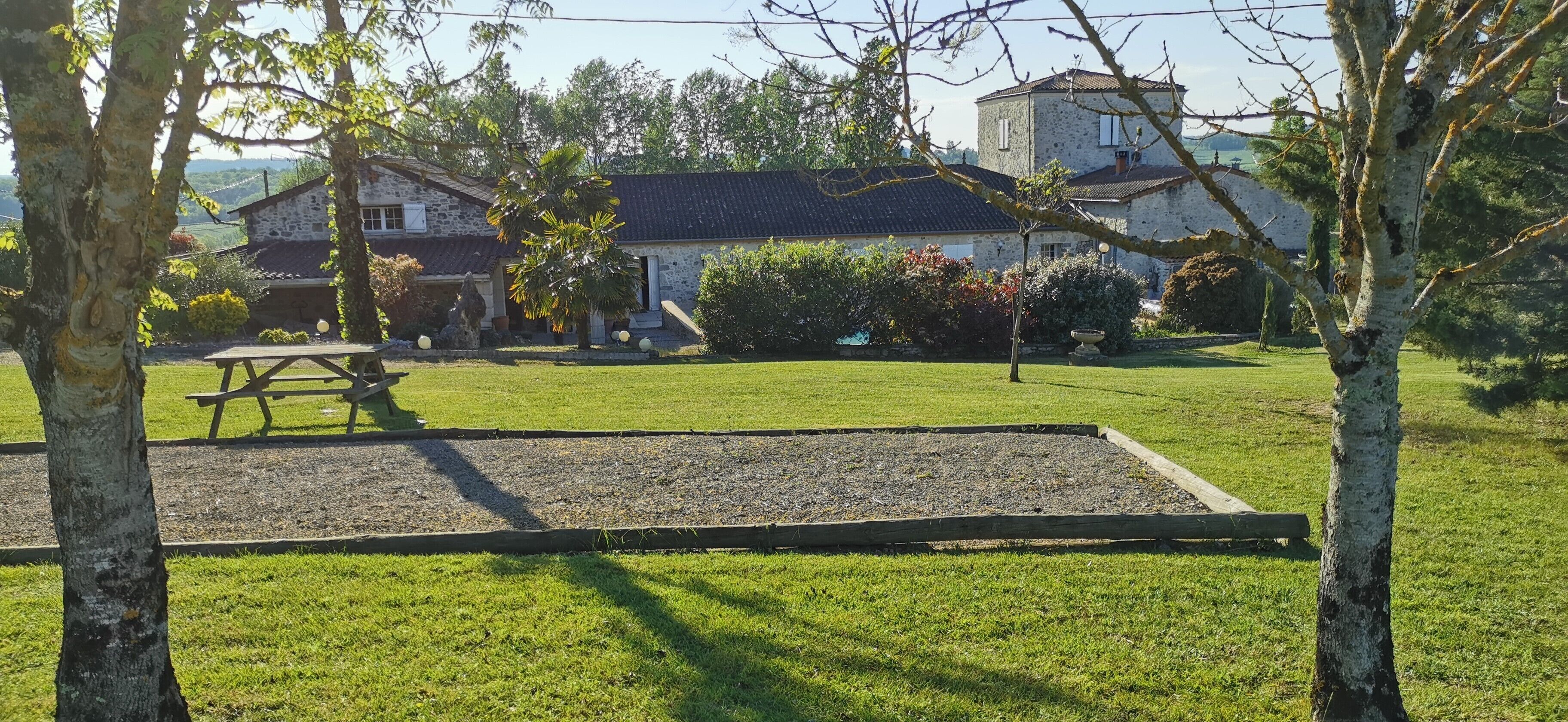 Une maison en pierre avec un toit en tuiles, entourée d’une pelouse bien entretenue et d’arbres.