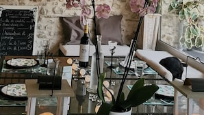 A cozy interior with a rustic stone wall, a wooden ceiling, a hanging wicker chandelier, a bookshelf with books and decorative items, a glass table with candles, and a window with plants.