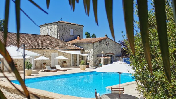A swimming pool with lounge chairs and umbrellas, surrounded by stone buildings with a tiled roof.