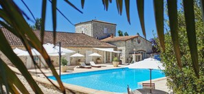 A swimming pool with lounge chairs and umbrellas, surrounded by stone buildings with a tiled roof.