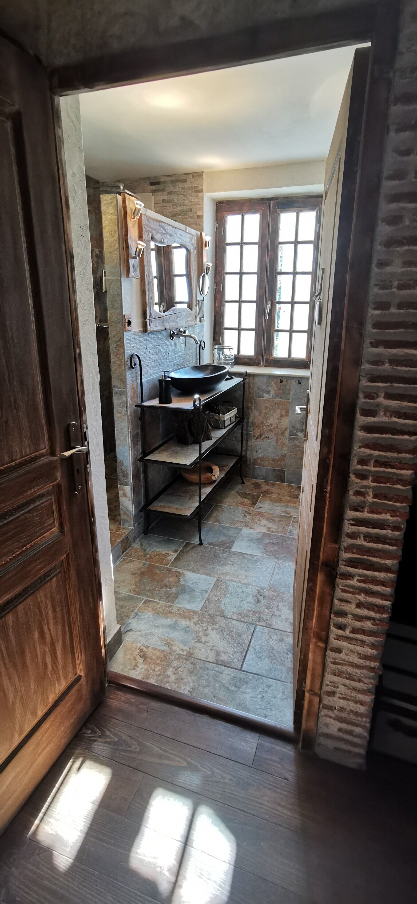 Une salle de bain avec un mur en pierre, un meuble-lavabo en bois, un lavabo noir et une fenêtre à carreaux.