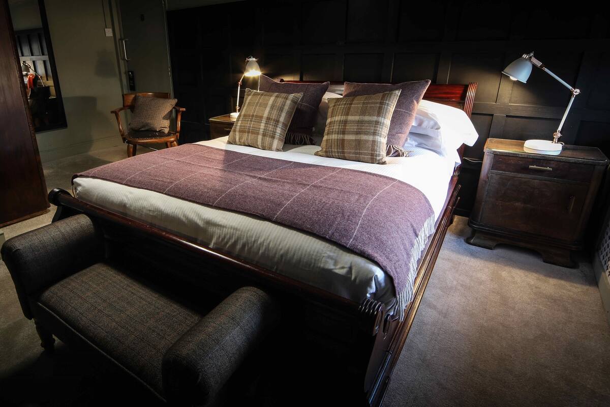 A neatly made bed with a headboard, two bedside lamps, a nightstand with a lamp, and a bench at the foot of the bed.