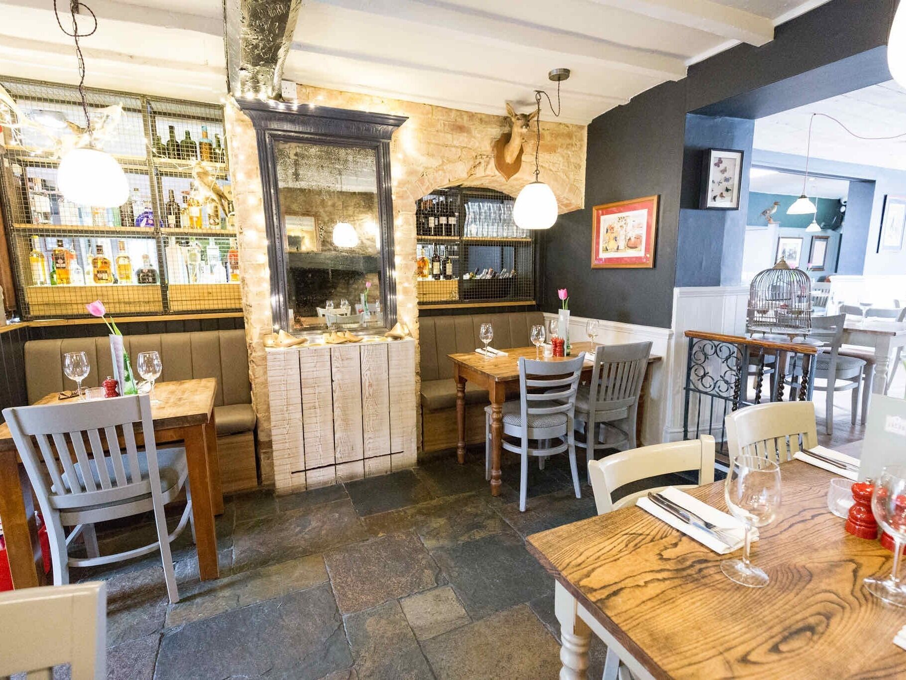 A restaurant interior with wooden tables and chairs, a bar area with bottles, and a stone wall.