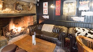 L’intérieur chaleureux d’un pub typique, avec une cheminée en pierre, des canapés en cuir et divers panneaux publicitaires annonçant des parties de fléchettes et d’autres activités.