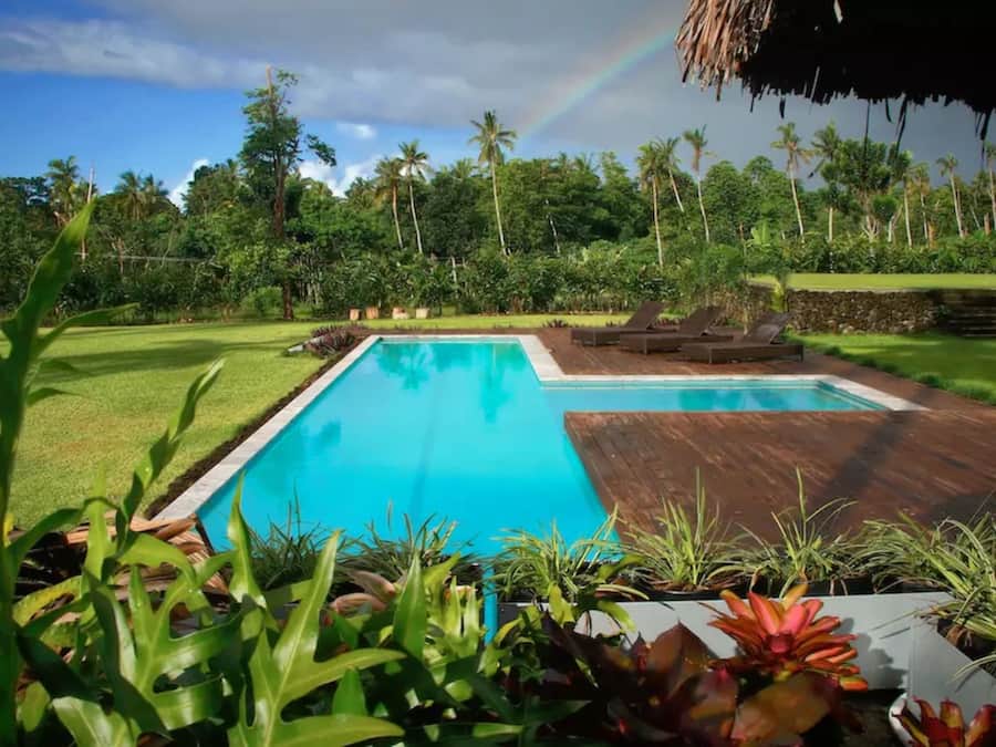 A swimming pool with a wooden deck, surrounded by lush greenery and palm trees.