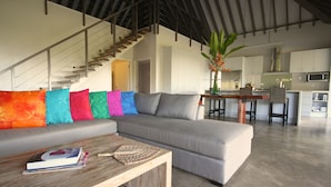 A modern living room with a grey sectional sofa, colorful cushions, a wooden coffee table with books, a staircase, and a kitchen in the background. - Ifiele'ele Plantation (Fasito'o Uta)