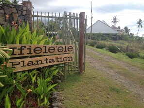 A wooden sign with the text "Ifiele'e Plantation" in front of a metal gate, surrounded by tropical plants and a stone wall. - Ifiele'ele Plantation (Fasito'o Uta)