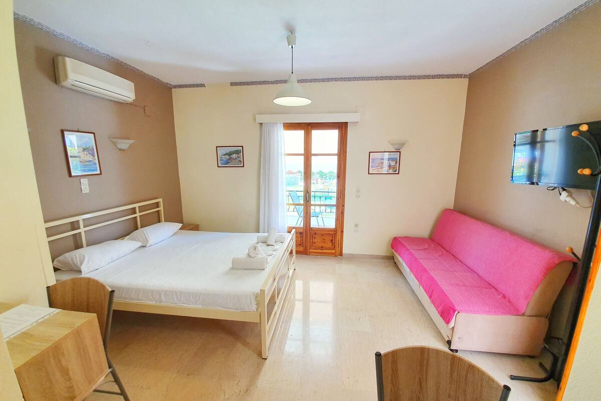 A hotel room with a bed, a desk, a chair, a sofa, a TV, and a window with a view of the outside.
