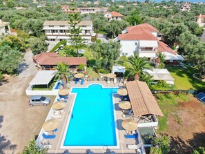 A large swimming pool surrounded by lounge chairs and umbrellas, with multiple buildings in the background. - Alexaria Holidays Apartments (Lefkada)