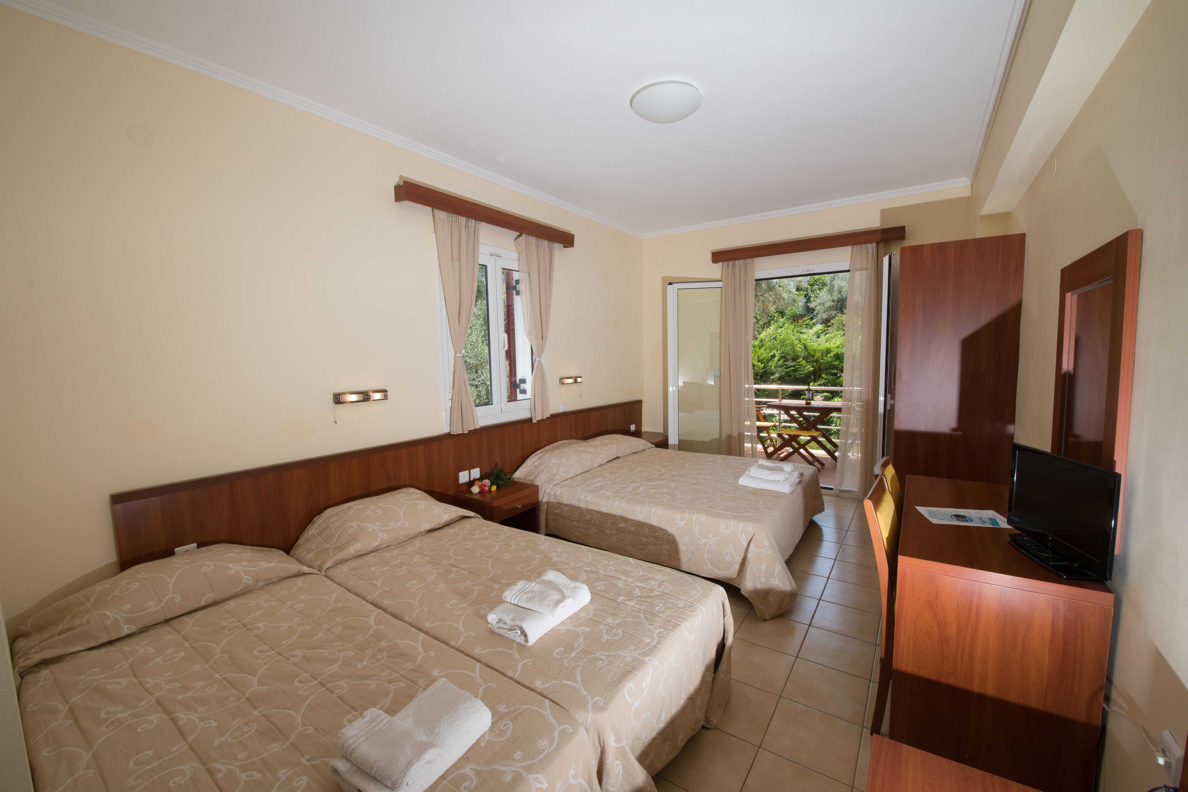 A hotel room with two beds, a desk with a television, and a balcony with a table and chairs.