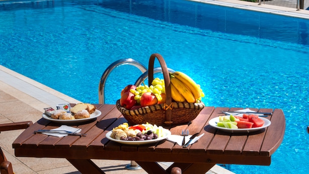 Preparación para un picnic junto a la piscina, con una mesa, platos de comida y una cesta de frutas.
