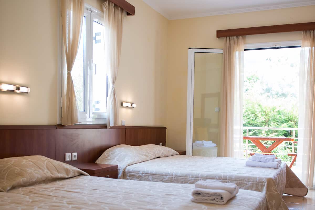 Ein Hotelzimmer mit zwei Betten, einem großen Fenster mit Vorhängen, einem Balkon und Blick ins Grüne.