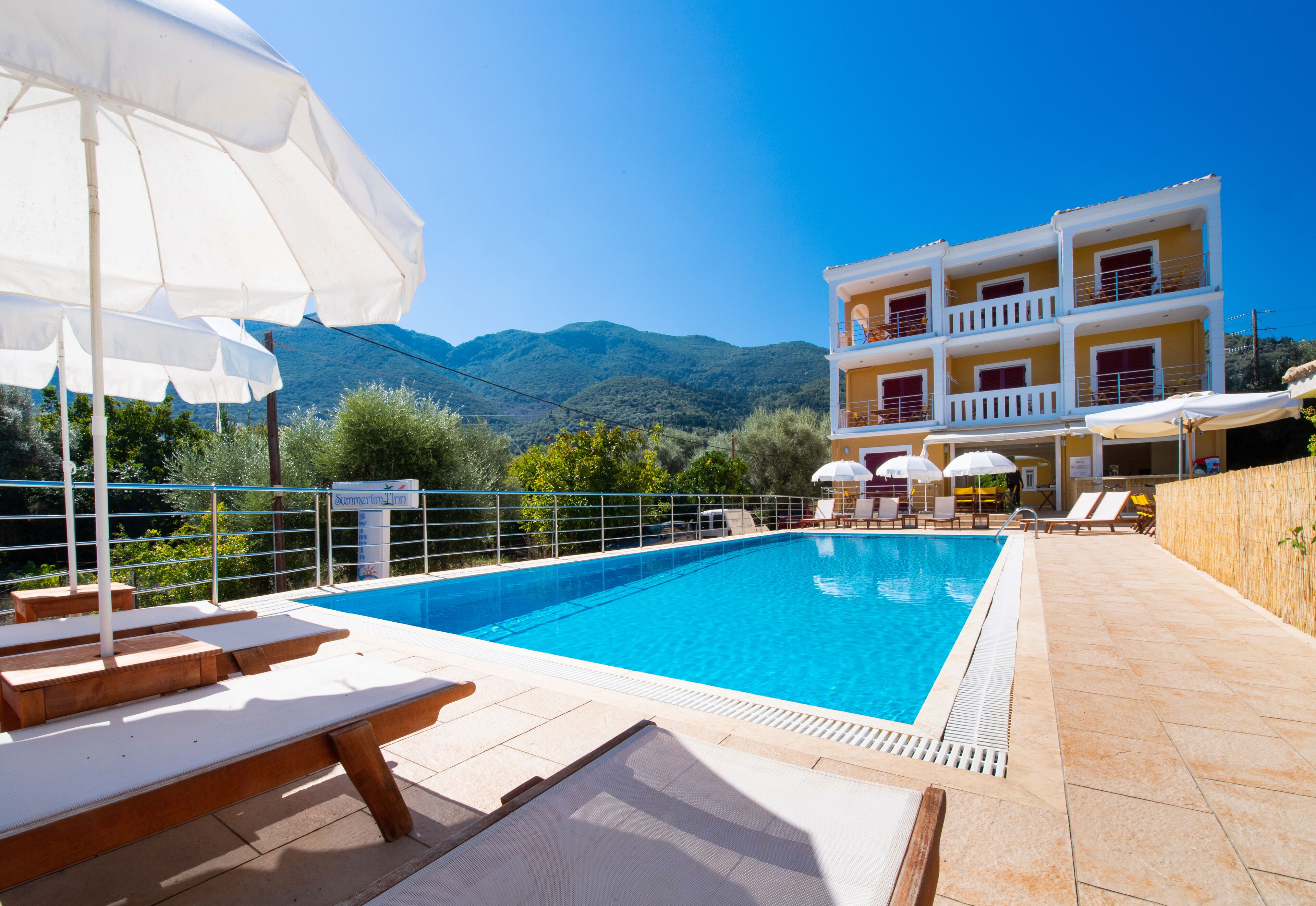 Piscina com espreguiçadeiras e guarda-sóis, um edifício com varandas e montanhas ao fundo.