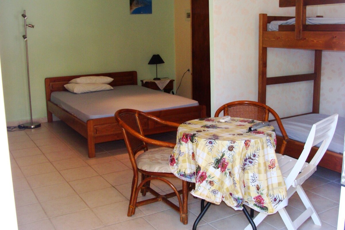 A bedroom with a single bed, a wooden chair, a small table with a floral tablecloth, and a bunk bed in the corner.