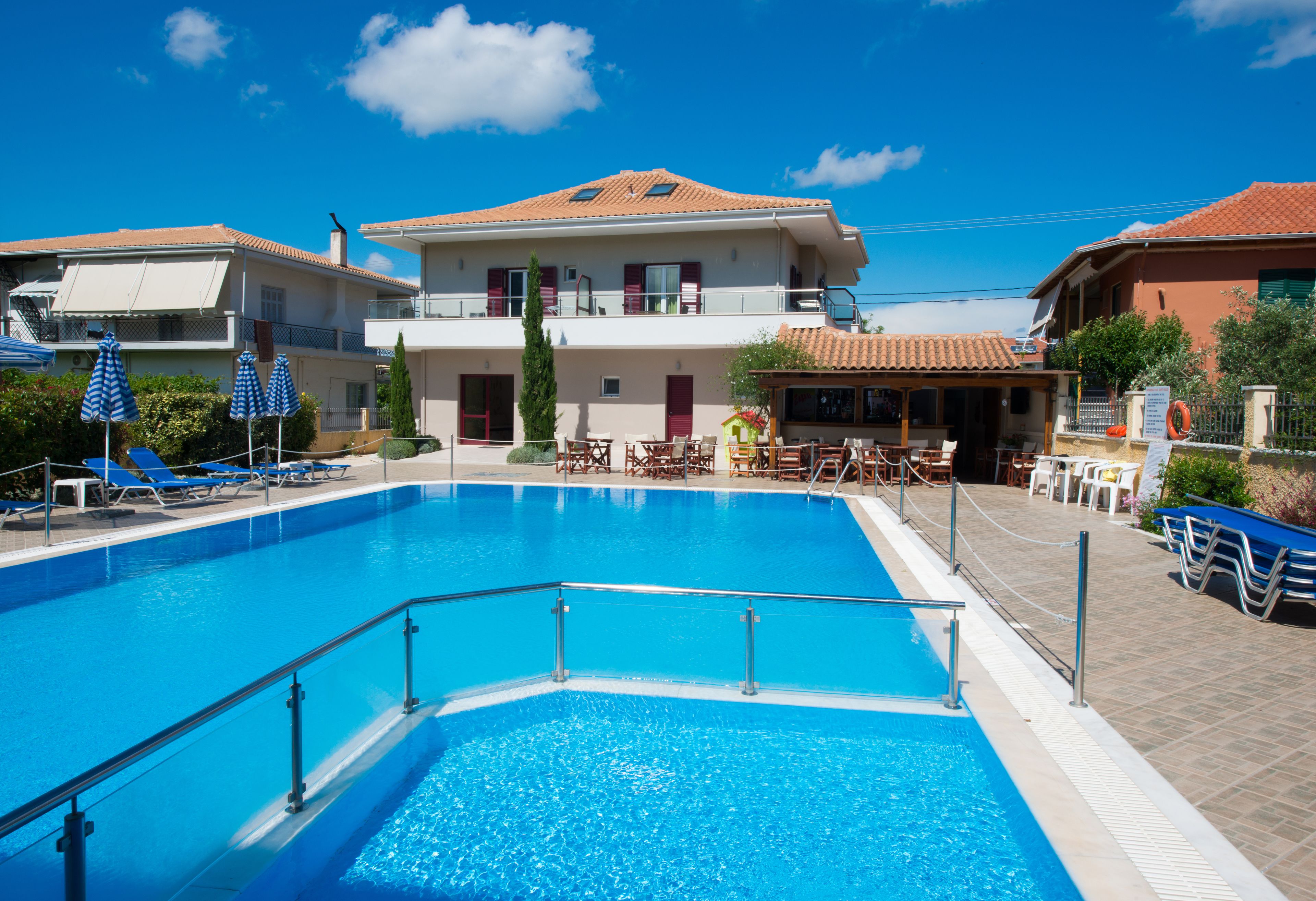 A swimming pool with lounge chairs and umbrellas, a building with a tiled roof, and a clear blue sky.