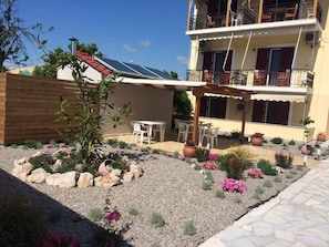 A two-story building with a balcony, a covered patio area with outdoor seating, and a garden with various plants and flowers. - Christina Studios (Lefkada)