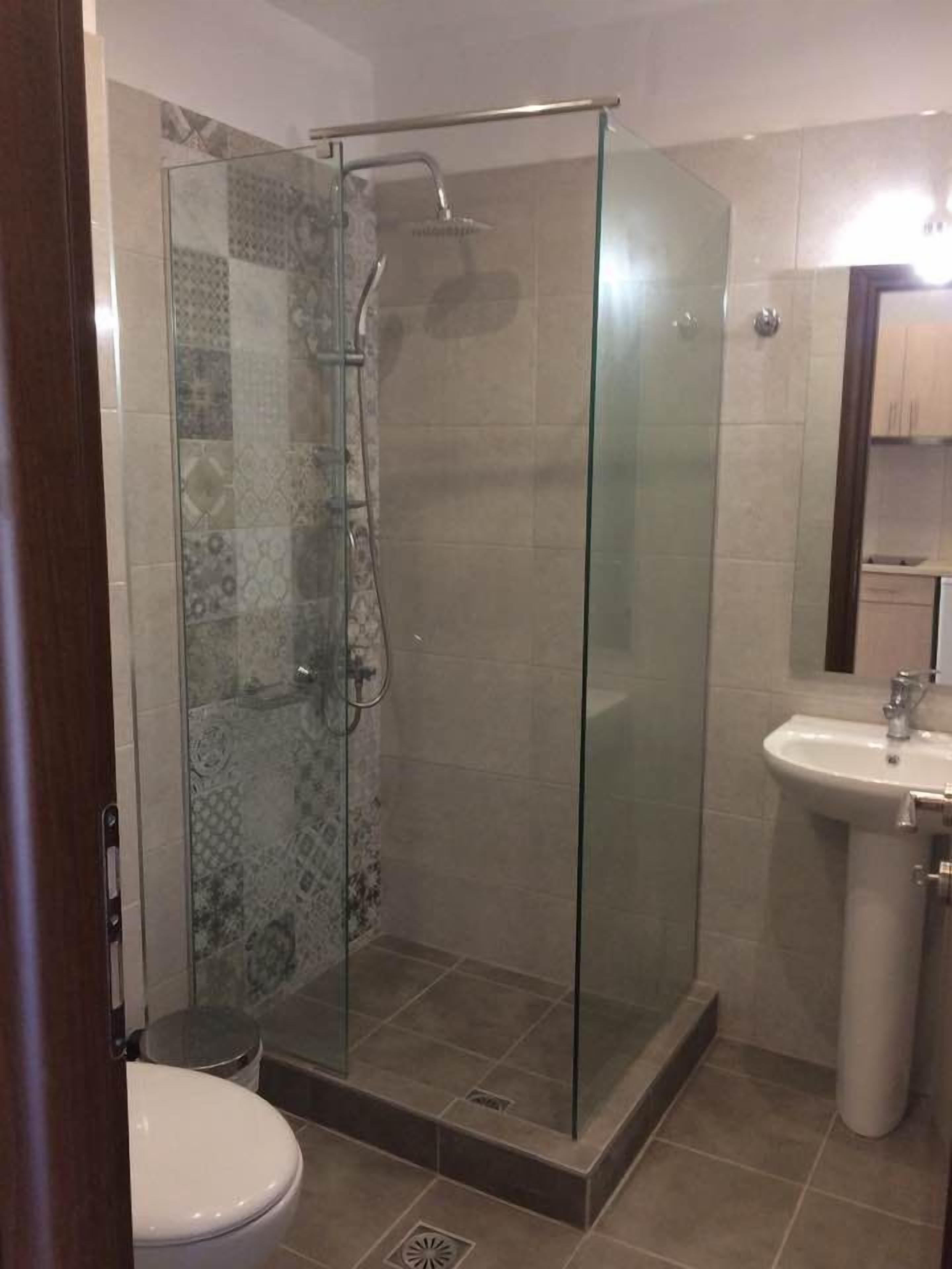 A glass-enclosed shower with a handheld showerhead, a white sink with a mirror above it, and a toilet with a patterned tile backsplash.