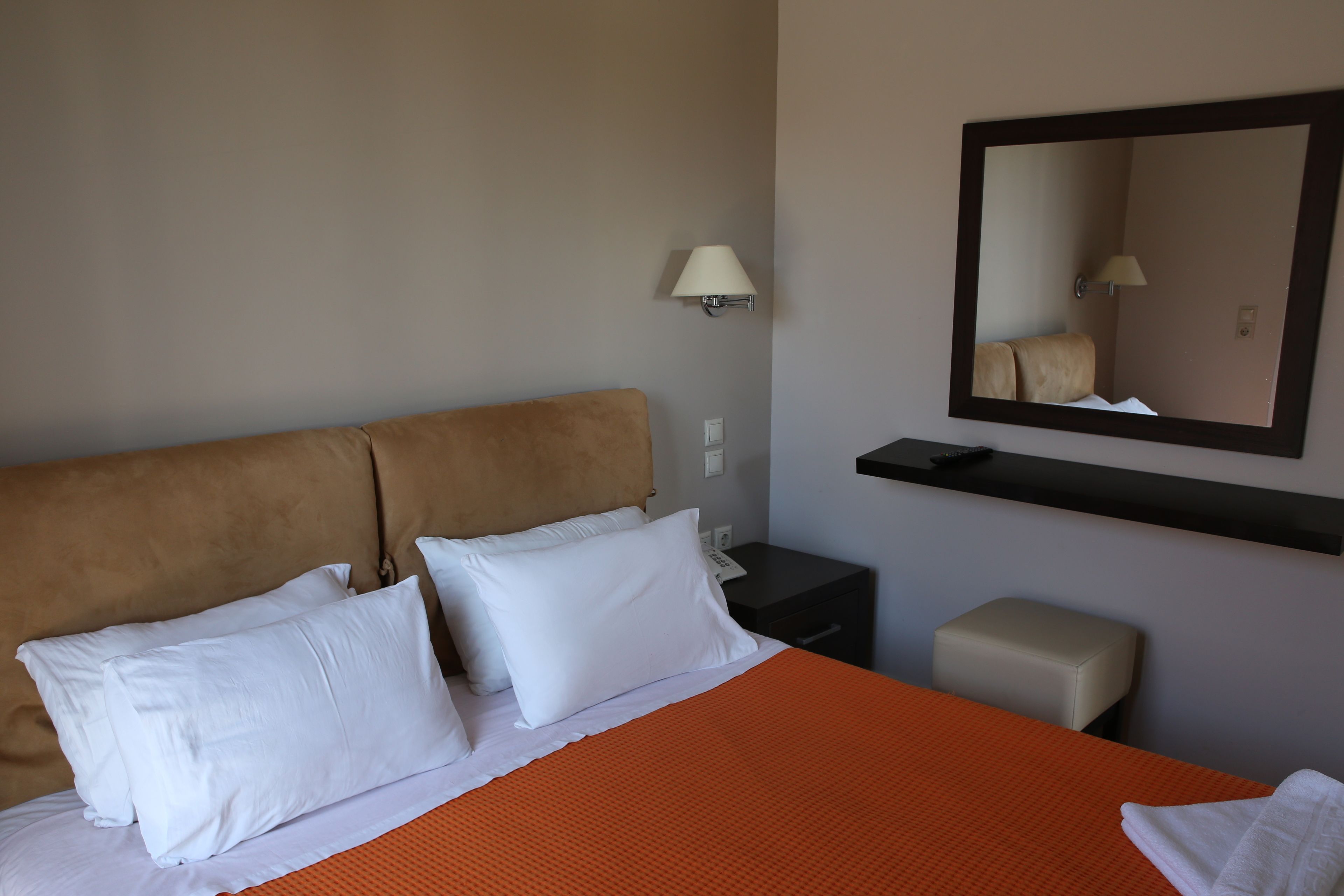 Een hotelkamer met een bed, kussens, een nachtkastje, een lamp, een spiegel en een klein krukje.