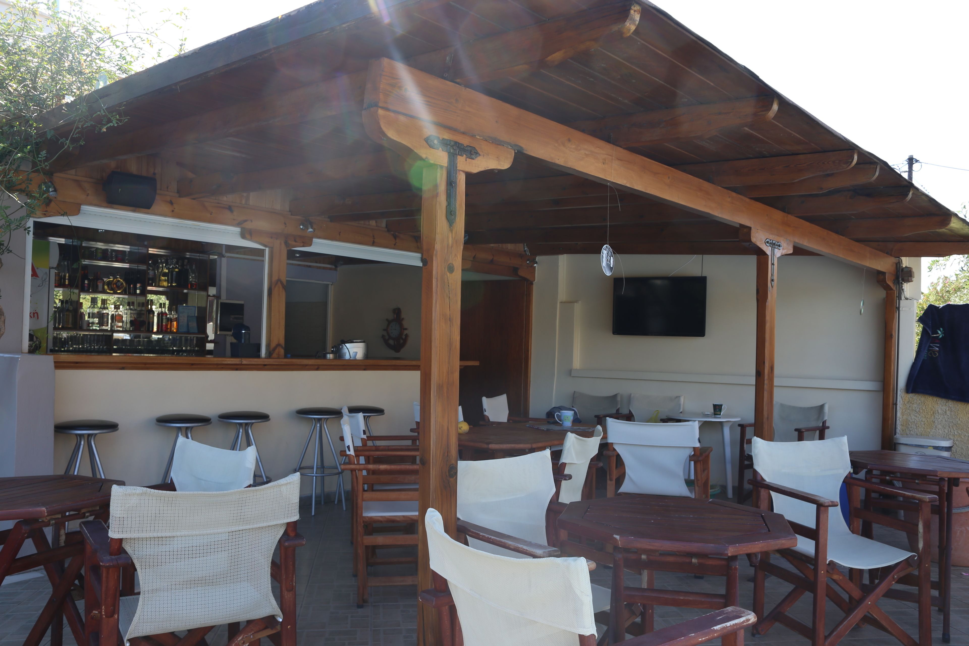 Ein Außenbereich mit einer Terrasse, Holztische und Stühle, eine Bar mit Hockern und ein an der Wand befestigter Fernseher.