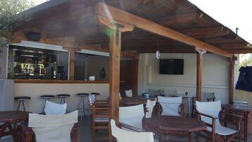 Ein Außenbereich mit einer Terrasse, Holztische und Stühle, eine Bar mit Hockern und ein an der Wand befestigter Fernseher.
