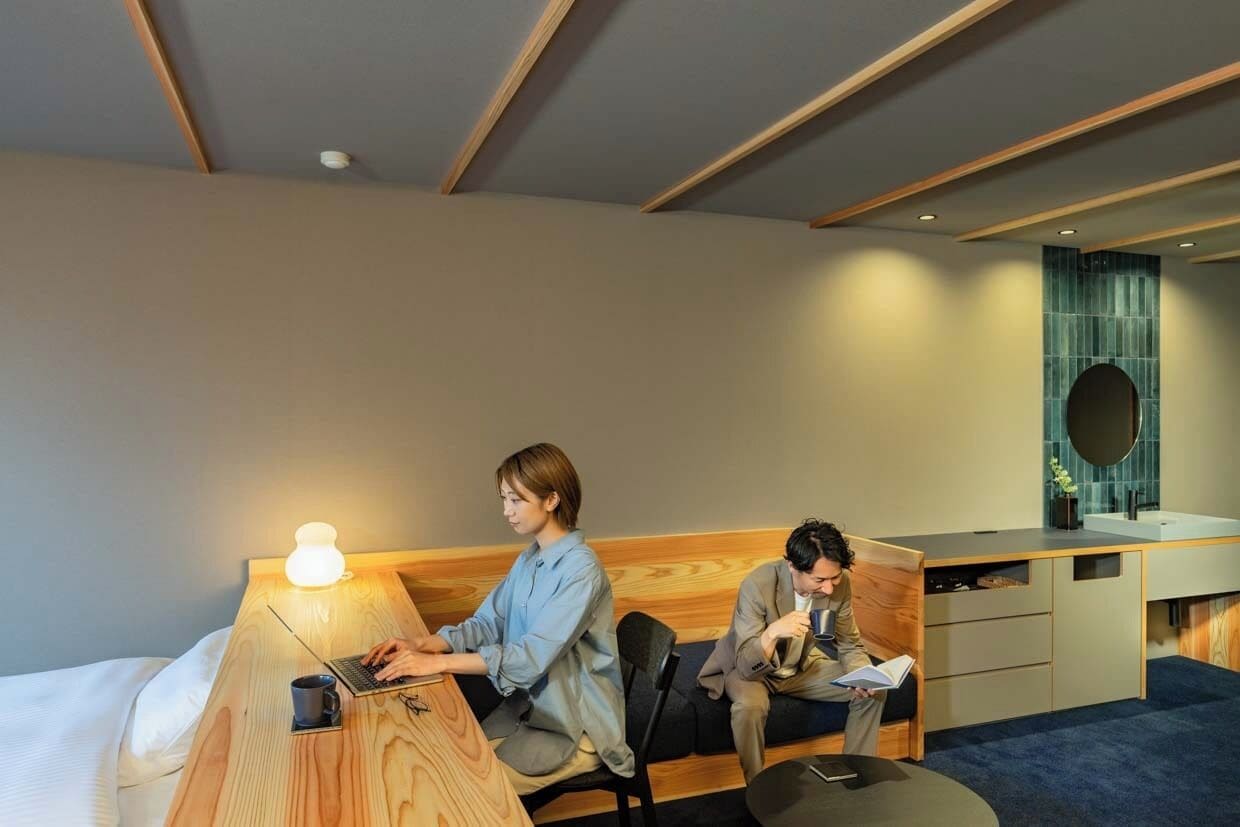 A modern hotel room with a wooden headboard, a desk with a laptop, a bedside table with a lamp, and a small round table with a chair.