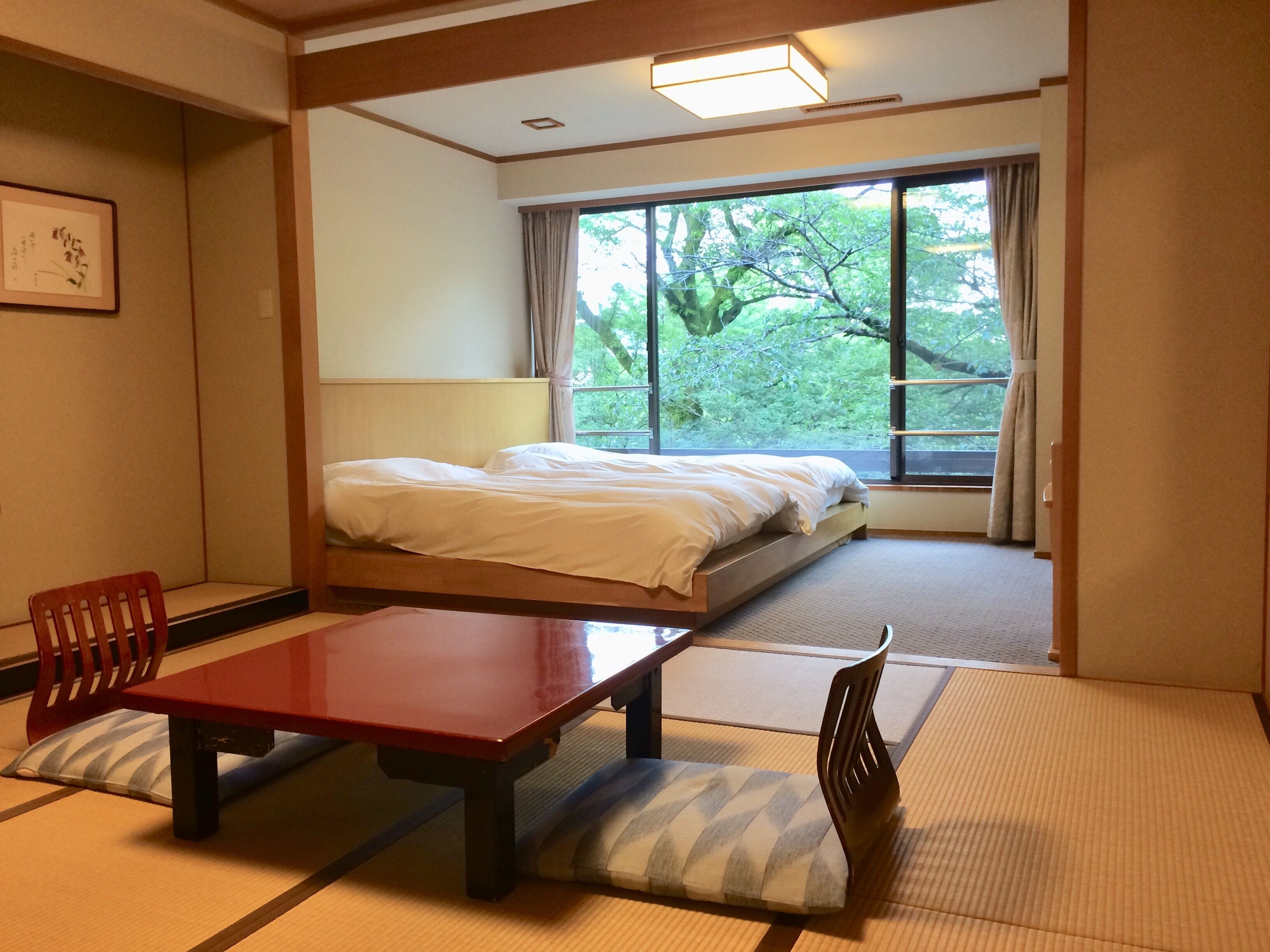 Une chambre d’hôtel avec un lit, une table en bois, une chaise, des tatamis et une grande fenêtre donnant sur des arbres.