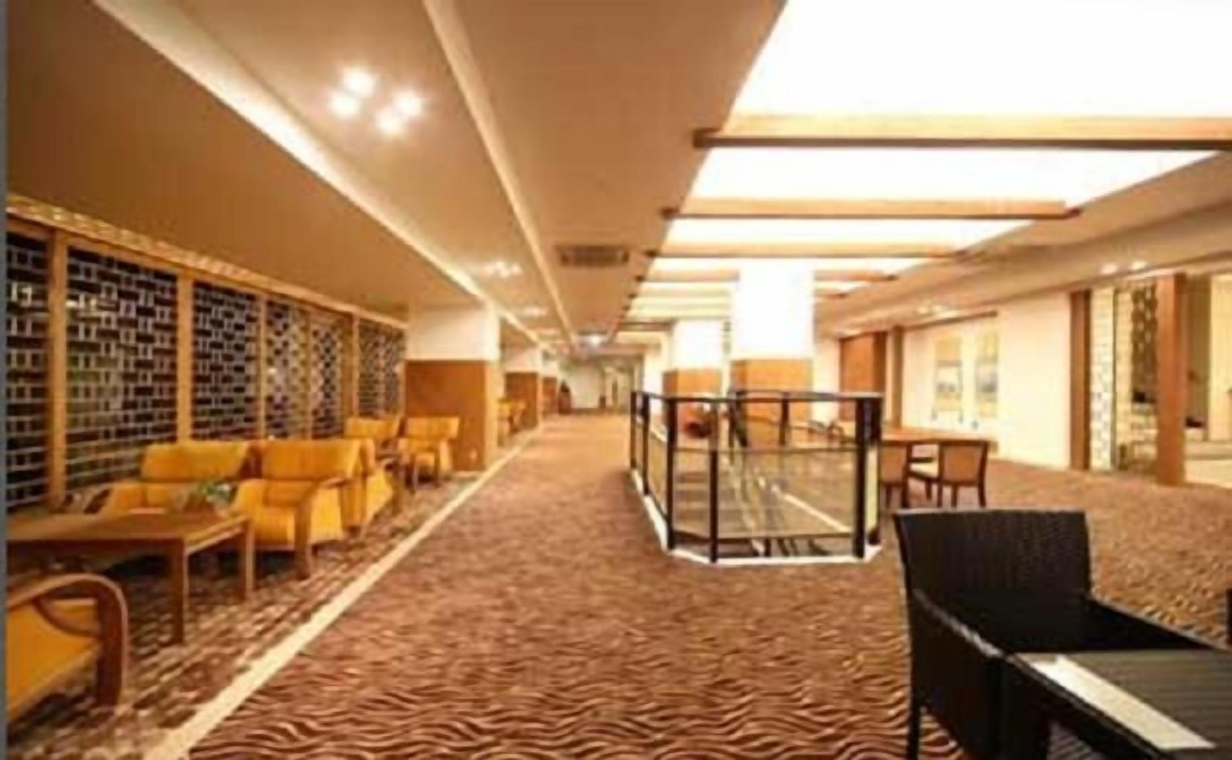A hotel lobby with a patterned carpet, seating area with yellow chairs, a glass partition, and a wall with a patterned design.