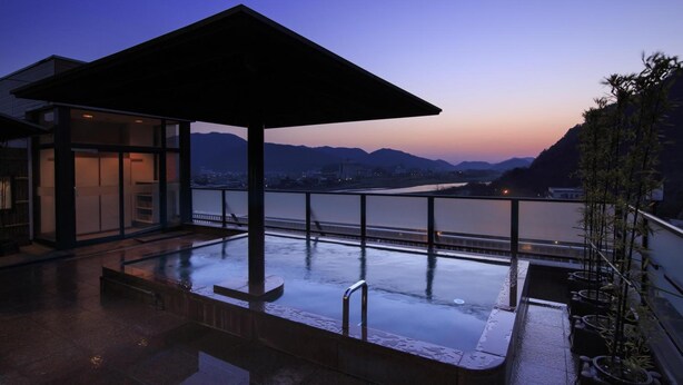 A modern outdoor pool area with a glass enclosure, a covered seating area, and a view of a river and mountains at dusk. - Hotel Park (Gifu)