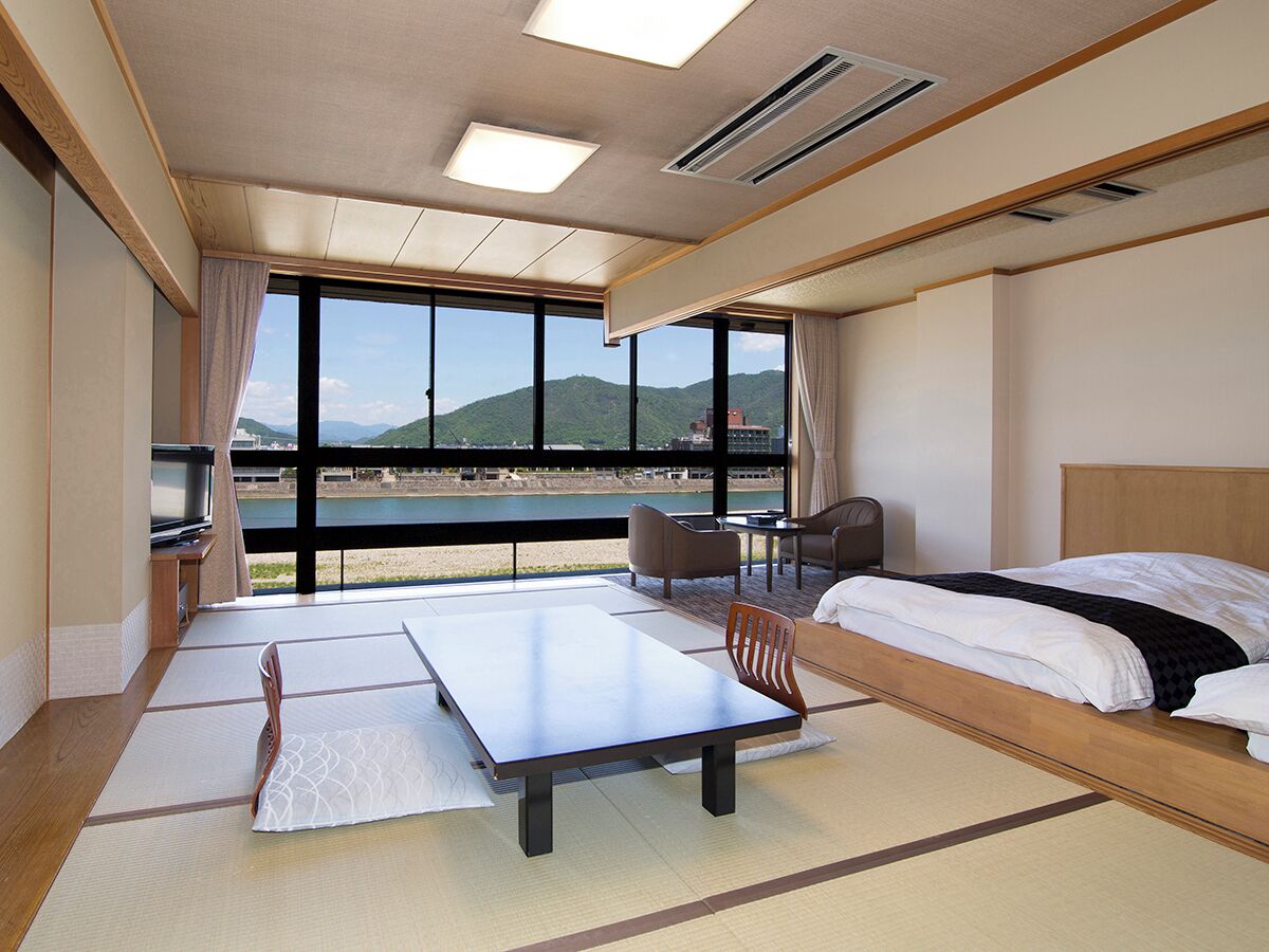 A hotel room with a bed, a table, a chair, a TV, and large windows offering a view of the outdoors.