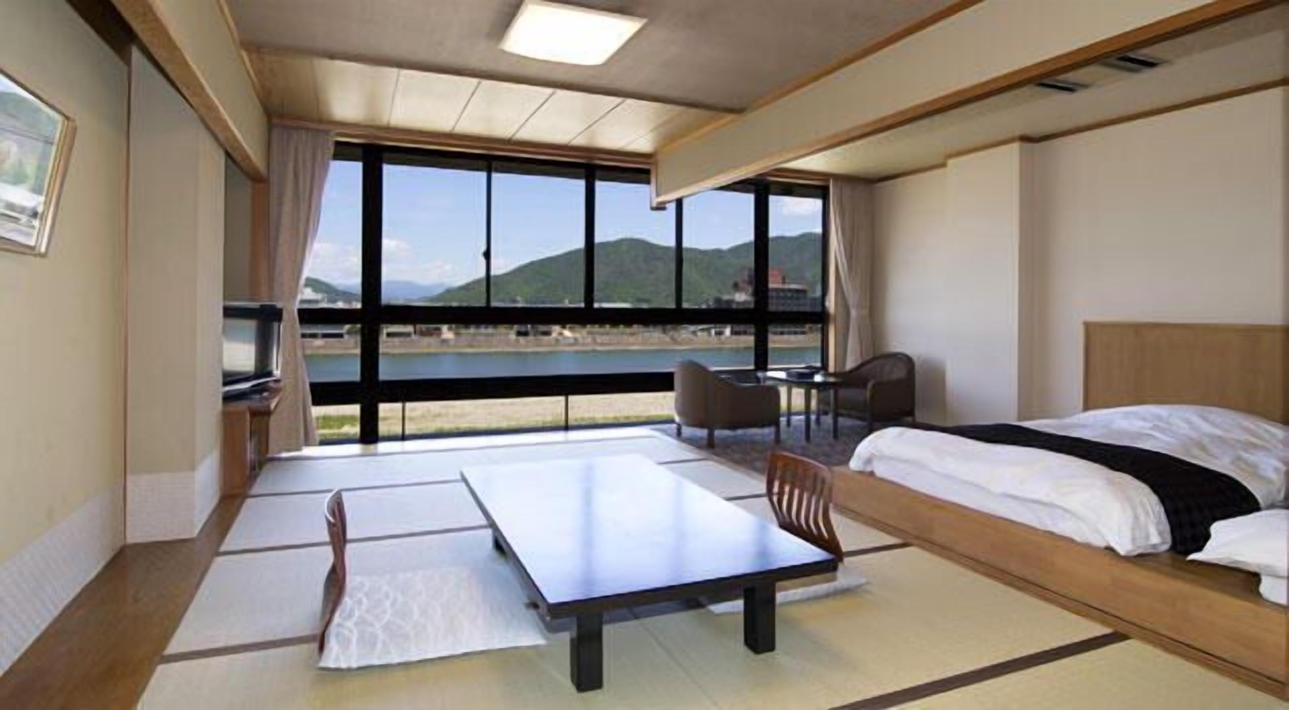 A hotel room with a bed, a desk, and a chair. There is a large window providing a view of the outdoors.