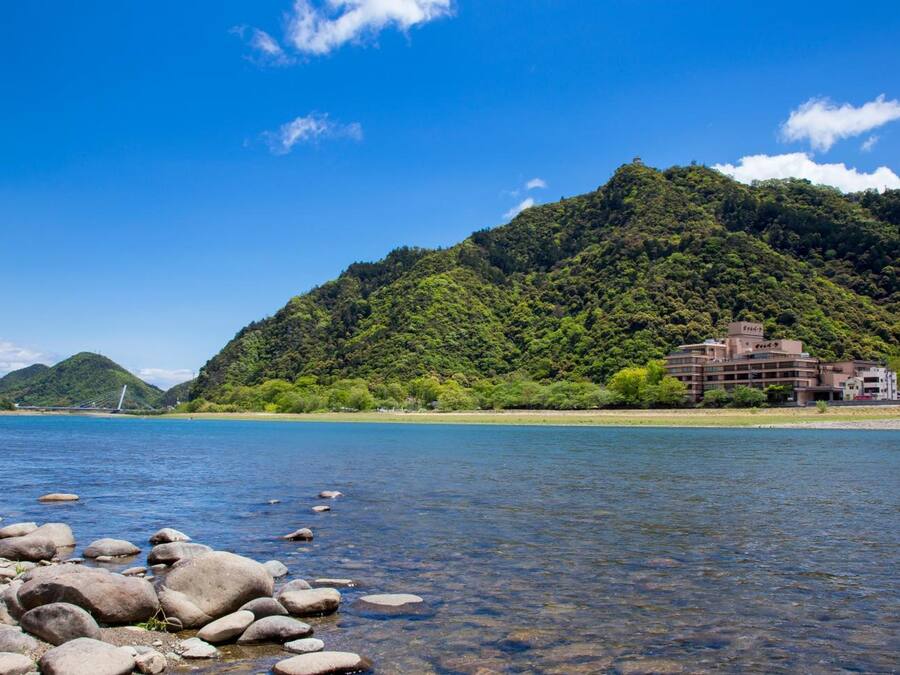 Un hotel costero con vista a un lago azul cristalino, una costa rocosa y una colina verde exuberante al fondo.
