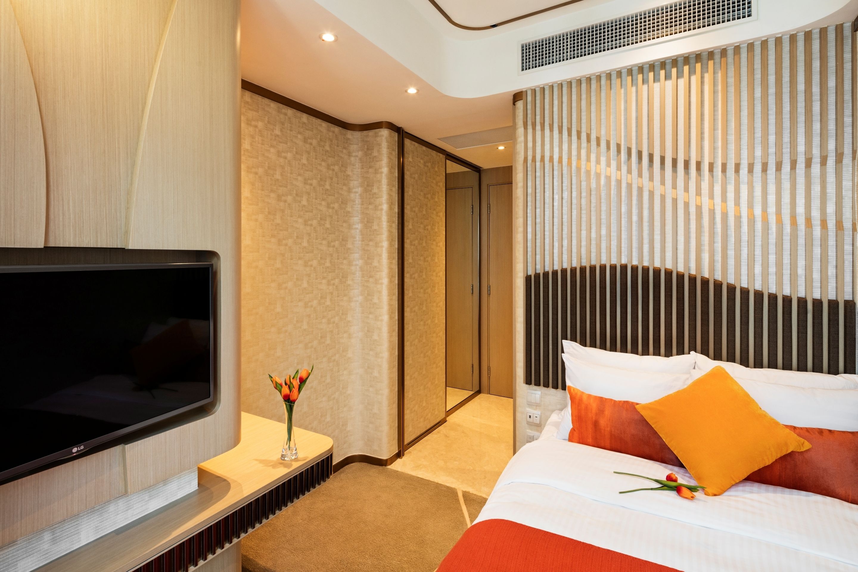 A modern hotel room with a large flat-screen TV, a bedside table with a vase of flowers, a bed with orange and white bedding, and a large mirror.