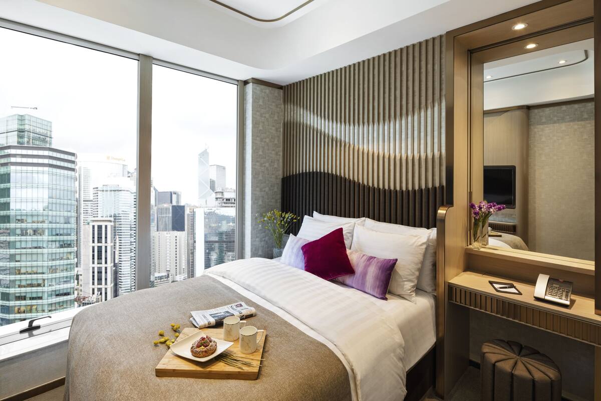 A modern hotel room with a large bed, a bedside table with a phone and a vase of flowers, a city view through large windows, and a cityscape visible outside.