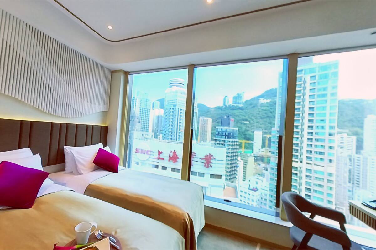A hotel room with a large bed, two pillows, a chair, and a view of a cityscape through large windows.